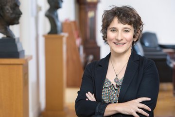 Auf dem Bild ist eIn vermutlich weibliches Exemplar der Gattung Homo sapiens in einem leicht verschwommen aufgenommenen Raum zu erkennen. Es trägt das dunkelblonde halblange Haar offen, lächelt freundlich und empathisch dem Betrachter entgegen, dabei die Arme verschränkt unterhalb der Brust gehalten. Die dunkle Jacke lässt unterhalb des Halses genügend Raum für den vollständig sichtbaren Kettenschmuck. Links im Bilde befinden sich auf Podesten mindestens zwei Büsten, die näher zu beschreiben nicht möglich ist.