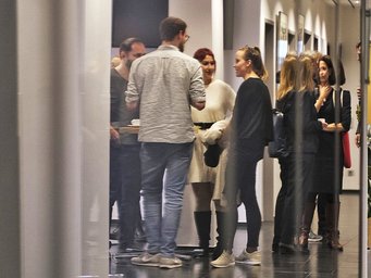 The image shows several specimens of the species Homo sapiens intellectualis standing in an informal circle in the entrance area of the institute. On the left side of the image, a curtain can be seen, and the view of the circle is somewhat obscured by mirror-like distortions, making the individuals appear slightly blurred.