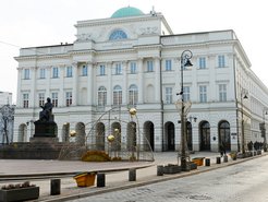 Auf dem Foto ist ein weißes Gebäude mit grüner Kuppel, Statue eines sitzenden Mannes und weihnachtlichen Dekorationen auf einem leeren Platz zu sehen. Es handelt sich um die Universität Warschau in Polen.