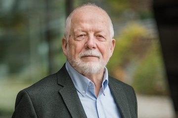 Mann trägt dunklen Anzug und hellblaues Hemd, Hintergrund zeigt unscharfe Herbstbäume, moderne Glasarchitektur.