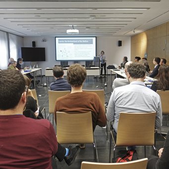 Konferenz Politik von Wachstum und Stagnation Das Foto gewährt Einblick in einen Konferenzraum, in dem Exemplare der Gattung Homo sapiens intellectualis an rechteckig angeordneten Tischen sitzend der Veranstaltung folgen. Im Hintergrund ist ein überdimensionaler Monitor zu erblicken, der vermutlich Präsentationszwecken des leitenden Individuums dient.