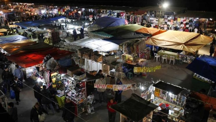 A vibrant night market with numerous stalls covered by colorful tents. Shoppers explore the diverse offerings under artificial lighting, creating a lively atmosphere.