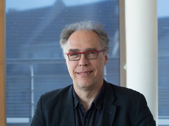 Akos Rona-Tas, 2018 als Scholar in Residence zu Gast am MPIfG Ein Mann in einem schwarzen Sakko sitzt vor einem Fenster, durch das unscharf Dächer und ein leicht bewölkter Himmel zu sehen sind.