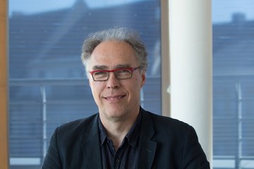 Ein Mann in einem schwarzen Sakko sitzt vor einem Fenster, durch das unscharf Dächer und ein leicht bewölkter Himmel zu sehen sind.