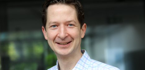 Ein Mann trägt ein blau-weiß kariertes Hemd, im Hintergrund sind unscharfe Fenster und Pflanzen zu sehen.