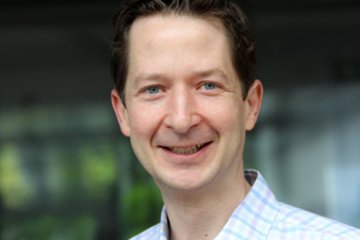 Ein Mann trägt ein blau-weiß kariertes Hemd, im Hintergrund sind unscharfe Fenster und Pflanzen zu sehen.