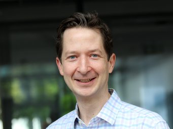 Tim Bartley, Scholar in Residence am MPIfG Ein Mann trägt ein blau-weiß kariertes Hemd, im Hintergrund sind unscharfe Fenster und Pflanzen zu sehen.