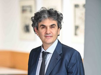 Lucio Baccaro, Scholar in Residence am MPIfG Person in einem dunkelblauen Anzug mit gepunkteter Krawatte, stehend in einem modernen, unscharfen Innenraum mit hellen und dunklen Flächen.