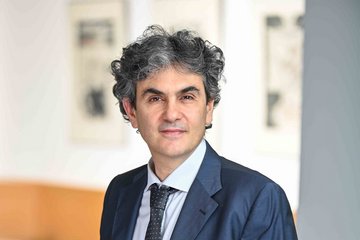 Person in einem dunkelblauen Anzug mit gepunkteter Krawatte, stehend in einem modernen, unscharfen Innenraum mit hellen und dunklen Flächen.