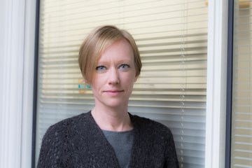 Eine Person steht vor einem Fenster mit geschlossenen Jalousien, gekleidet in einem grauen Shirt und einem dunklen, texturierten Cardigan.