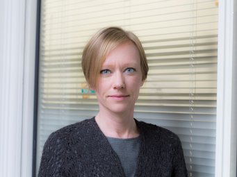 Jenny Andersson, Associate Fellow des Max Planck Sciences Po Center on Coping with Instability in Market Societies (MaxPo), Paris, und Assoziierte Wissenschaftlerin des MPIfG Eine Person steht vor einem Fenster mit geschlossenen Jalousien, gekleidet in einem grauen Shirt und einem dunklen, texturierten Cardigan.
