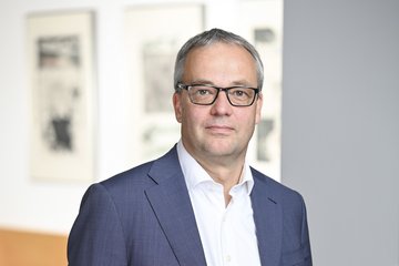 Ein Mann trägt einen blauen Anzug mit weißem Hemd und schwarzem Gürtel. Im Hintergrund sind moderne Kunstwerke und ein Holztisch zu sehen.