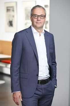 Ein Mann trägt einen blauen Anzug mit weißem Hemd und schwarzem Gürtel. Im Hintergrund sind moderne Kunstwerke und ein Holztisch zu sehen.