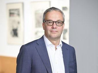 Jens Beckert, seit 2005 Direktor am MPIfG und Professor für Soziologie an der Wirtschafts- und Sozialwissenschaftlichen Fakultät der Universität zu Köln Ein Mann trägt einen blauen Anzug mit weißem Hemd und schwarzem Gürtel. Im Hintergrund sind moderne Kunstwerke und ein Holztisch zu sehen.