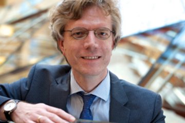 Ein Mann im Business-Anzug, mit einer silbernen Armbanduhr und einer blauen Krawatte, lehnt an einer modernen Glasarchitektur im Hintergrund.