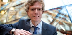 Ein Mann im Business-Anzug, mit einer silbernen Armbanduhr und einer blauen Krawatte, lehnt an einer modernen Glasarchitektur im Hintergrund.