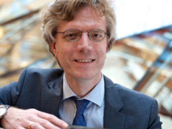 Werner Eichhorst, Vorsitzender des Vereins der Freunde und Ehemaligen des MPIfG Ein Mann im Business-Anzug, mit einer silbernen Armbanduhr und einer blauen Krawatte, lehnt an einer modernen Glasarchitektur im Hintergrund.