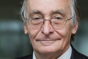 Graue Haare, eleganter Anzug mit weißem Hemd und farbiger Krawatte vor einem unscharfen Hintergrund mit Glasfenstern.