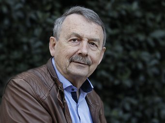 Wolfgang Streeck, Direktor am MPIfG Grauhäutiger Mann mit brauner Lederjacke und hellblauem Hemd steht vor einem Hintergrund aus unscharfem Laubwerk.