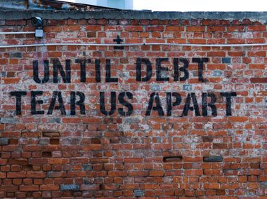 Eine Backsteinwand mit einem Graffiti-Slogan in schwarzen Buchstaben: „Until debt tear us apart“. Die Wand weist einige Verwitterungsspuren auf.