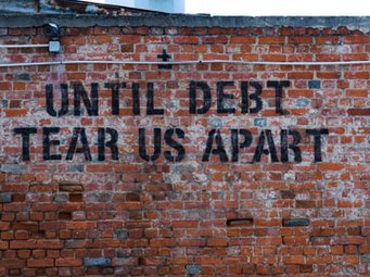 Eine Backsteinwand mit einem Graffiti-Slogan in schwarzen Buchstaben: „Until debt tear us apart“. Die Wand weist einige Verwitterungsspuren auf.