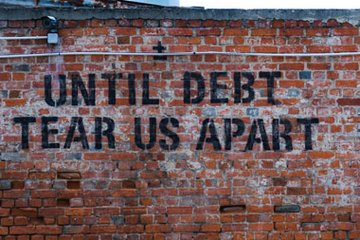Eine Backsteinwand mit einem Graffiti-Slogan in schwarzen Buchstaben: „Until debt tear us apart“. Die Wand weist einige Verwitterungsspuren auf.