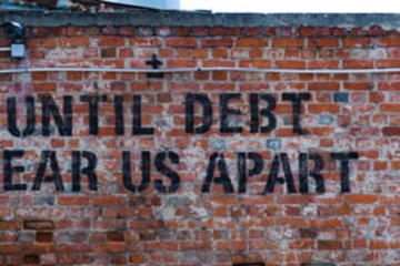 Auf einer roten Backsteinmauer ist ein schwarzer Schriftzug zu sehen, der den Spruch „Until debt tear us apart“ enthält. In der oberen Ecke ist eine Überwachungskamera angebracht.