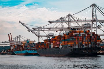 Im Vordergrund befinden sich zwei beladene Containerschiffe in einem Hafen. Mehrere Kräne erstrecken sich über die Schiffe, während im Hintergrund weitere Schiffe am Kai zu sehen sind.