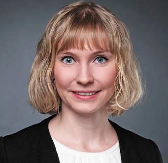 Marina Hübner Ein Exemplar der Gattung Homo sapiens intellectualis ist auf dem Foto zu bestaunen. Es hat schulterlange blonde Haare, das in Wellen herabhängt, und trägt eine schwarze Jacke mit weißer Bluse darunter. Die Szene ist vor einem dunkelgrauen Hintergrund situiert. Das Individuum hat sich ein schüchternes Lächeln ins Gesicht gezaubert und blickt aus den blauen, offenen Augen dem Betrachter entgegen.