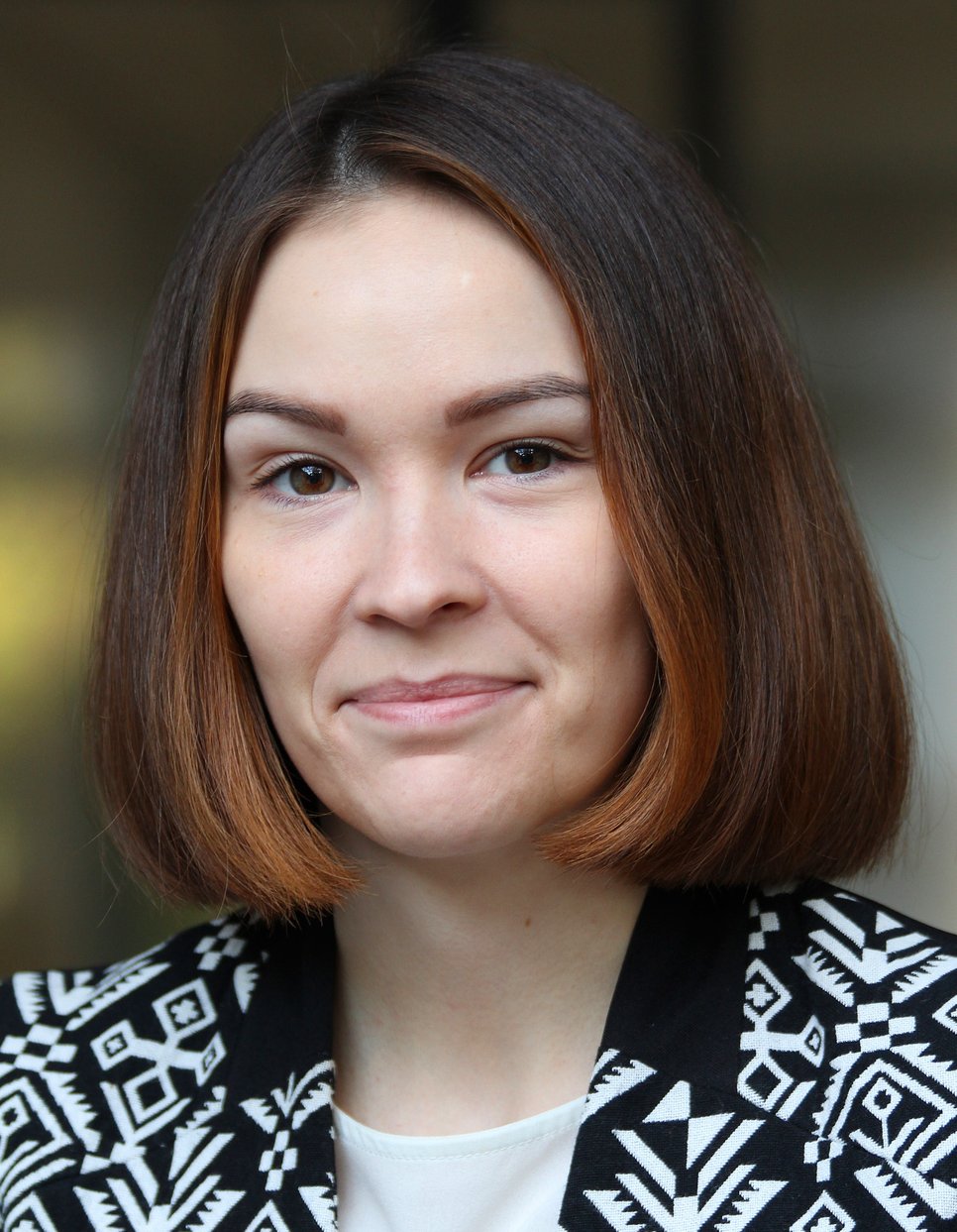 Eine Person mit schulterlangem, dunkelbraunen Haar mit einem Bob-Schnitt ist auf dem Foto zu erkennen. Die Jacke zeigt ein schwarz-weißes geometrisches Muster. Im Hintergrund sind unscharfe Details sichtbar.