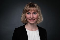 A specimen of the species Homo sapiens intellectualis can be admired in the photo. It has shoulder-length blonde hair that hangs down in waves and is wearing a black jacket with a white blouse underneath. The scene is set against a dark gray background. The individual has a shy smile on their face and looks at the viewer with blue, open eyes.