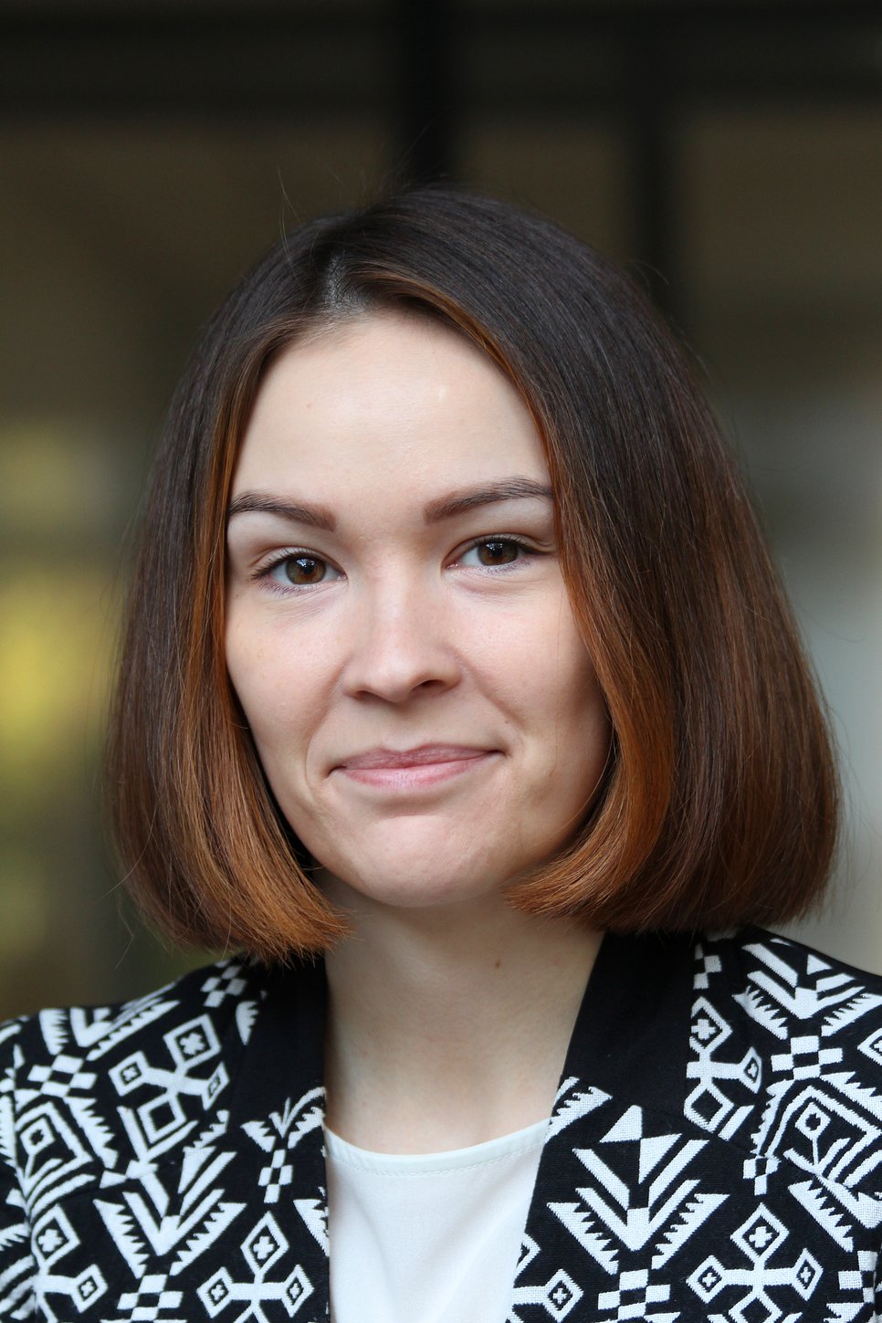 A person with shoulder-length, dark brown hair in a bob cut can be seen in the photo. The jacket has a black and white geometric pattern. Blurred details are visible in the background.