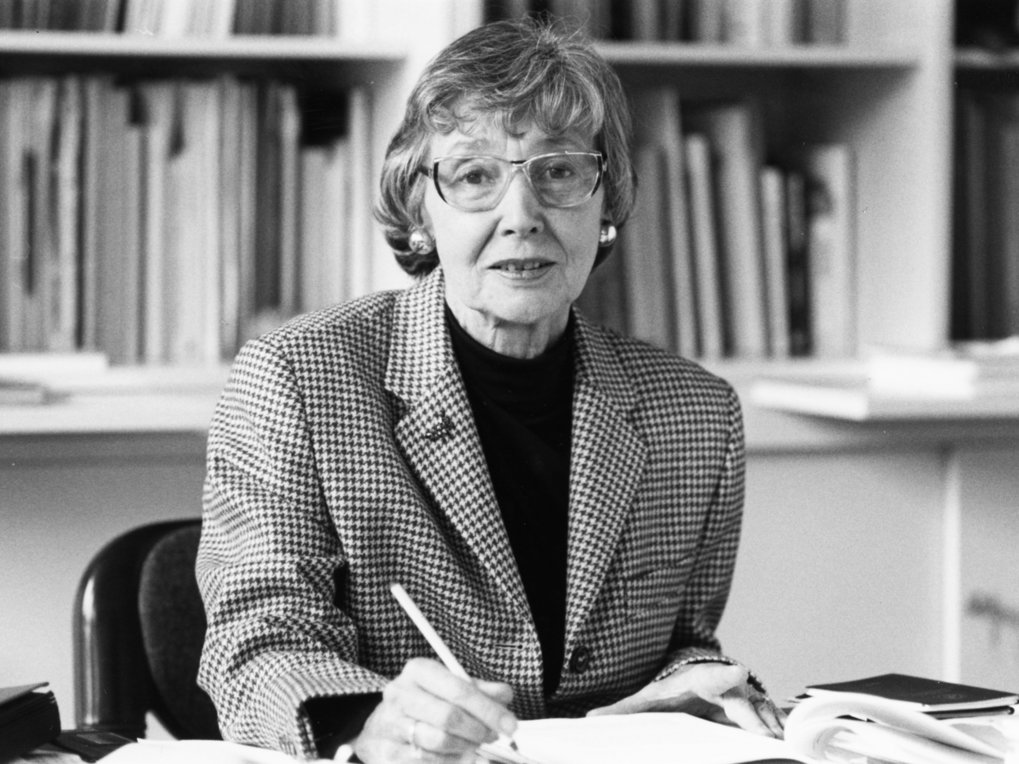 Eine Person in einer karierten Jacke sitzt an einem Tisch und schreibt in ein aufgeschlagenes Buch, während im Hintergrund ein Bücherregal mit zahlreichen Büchern zu sehen ist.