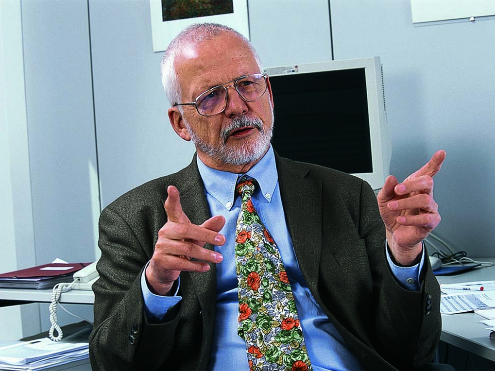 Eine Person in einem Bürosetting trägt ein dunkelgraues Sakko und eine bunte Krawatte mit Blumenmuster. Im Hintergrund sind ein Computer, Akten und ein Telefon sichtbar, was auf eine Arbeitsumgebung hinweist.