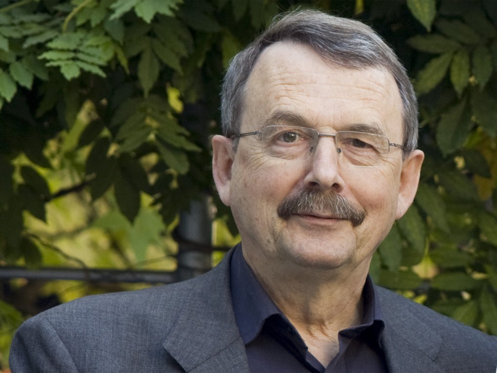 Person mit dunklem Blazer, im Freien mit dichtem Grün im Hintergrund, was auf eine natürliche Umgebung hindeutet.