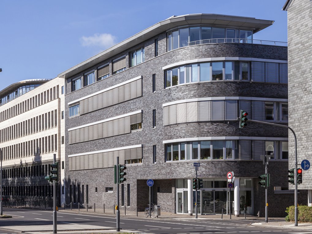 Ein modernes Gebäude mit geschwungener Fassade und großen Fenstern an einer städtischen Straßenecke, umgeben von Ampeln und einem Radweg. Es handelt sich um das Gebäude des Max-Planck-Instituts für Gesellschaftsforschung in Köln.