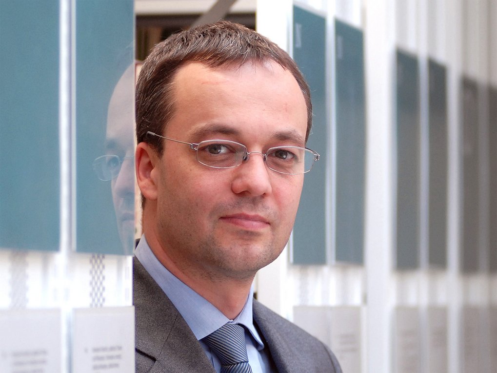 Ein Mann in einem grauen Anzug mit blauer Krawatte steht vor einer Reihe von vertikalen Informationstafeln, die in einem modernen Gebäude platziert sind.