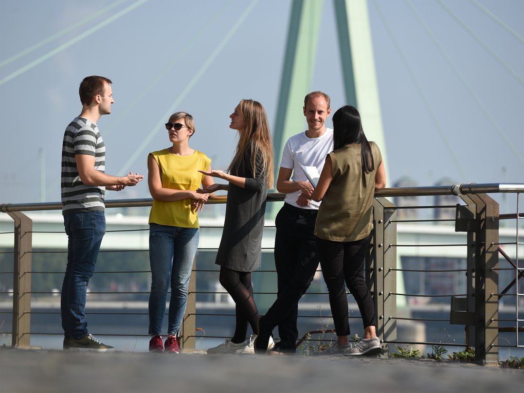 Fünf Personen stehen auf einer Uferpromenade und unterhalten sich. Hinter ihnen überspannt eine große Brücke den Fluss unter klarem Himmel.