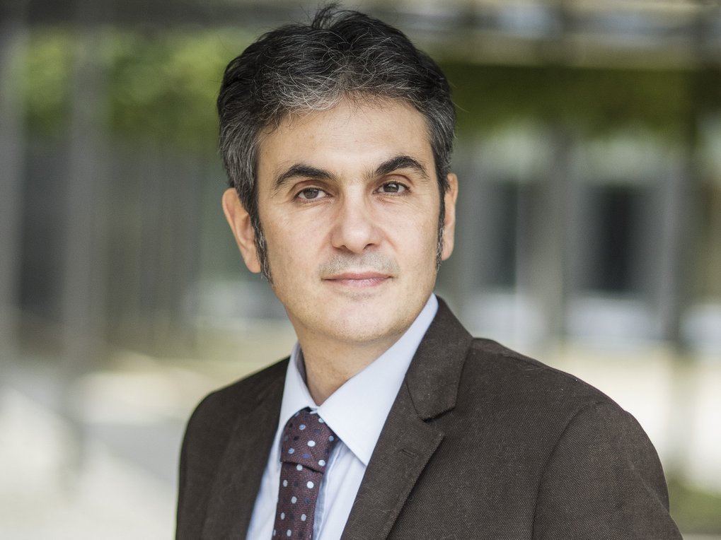 Ein Mann trägt einen dunkelbraunen Anzug mit weißem Hemd und eine dunkelrote Krawatte mit weißen Punkten. Der Hintergrund ist verschwommen und zeigt ein Gebäude mit Glasfronten und grüner Vegetation.