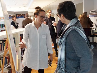 Posterpräsentation im Rahmen einer Evaluation des MPIfG Zwei Personen unterhalten sich angeregt vor einer Präsentationstafel in einer gut besuchten Bibliothek. Im Hintergrund sind Bücherregale, weitere Präsentationstafeln und Personen in Gesprächen zu sehen.