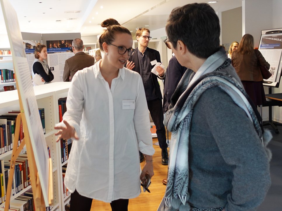 Zwei Personen unterhalten sich angeregt vor einer Präsentationstafel in einer gut besuchten Bibliothek. Im Hintergrund sind Bücherregale, weitere Präsentationstafeln und Personen in Gesprächen zu sehen.