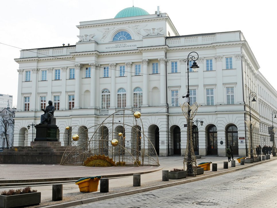 Ein großes neoklassizistisches Gebäude mit einer grünen Kuppel, dekorativen Straßenlaternen und einer Statue einer sitzenden Figur auf einem Sockel davor.