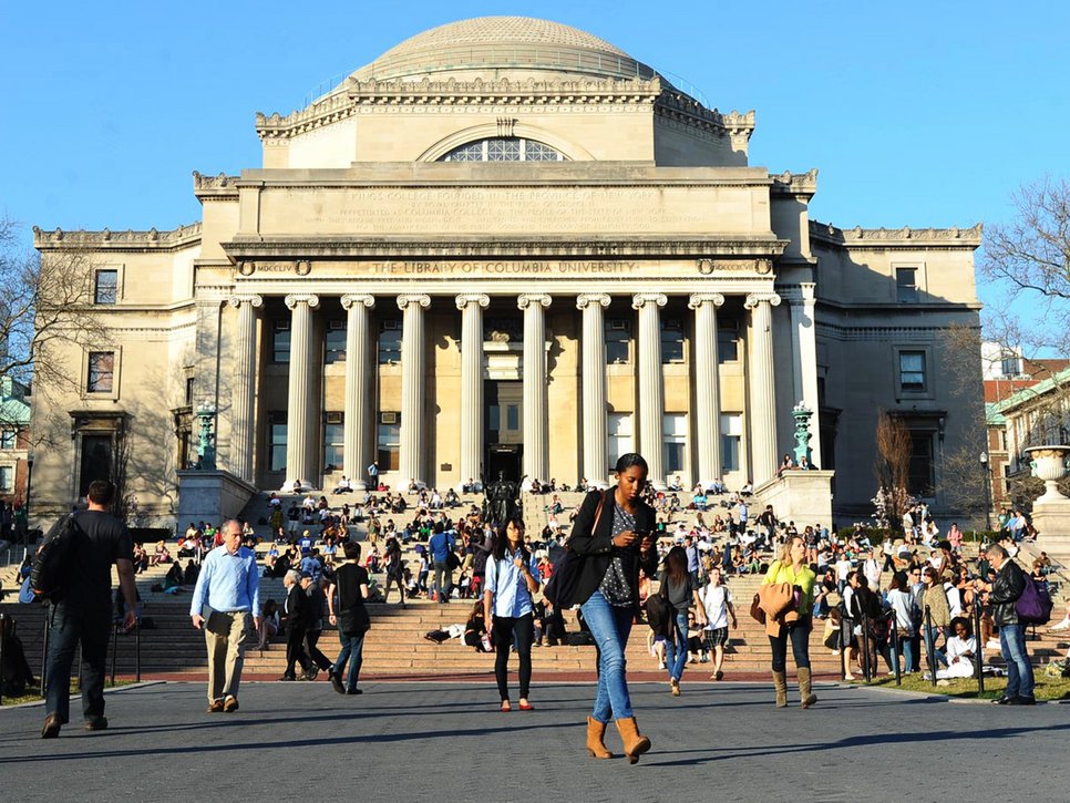Eine Gruppe von Menschen versammelt sich auf den breiten Stufen der Columbia University Library, während einige sich entspannen und andere vorbeigehen.