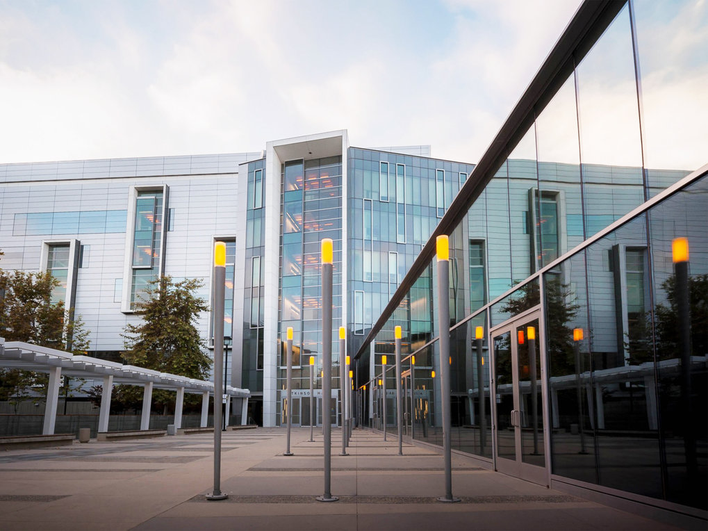 Large glass building facade with reflective windows, vertical yellow light bars in the entrance area, and surrounding trees.