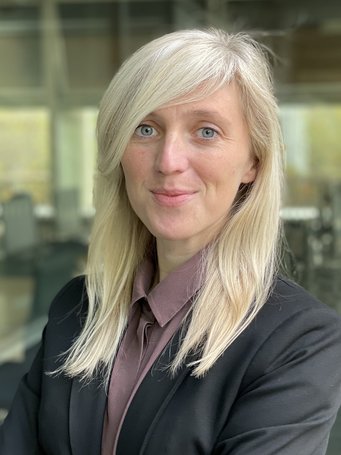 Sarah Berens ist Assistenz-Professorin an der Universität Innsbruck Person mit blonden Haaren, schwarzem Sakko und lila Bluse vor einem verschwommenen Bürohintergrund.