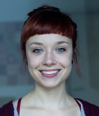 Laura Einhorn am Institut für Sozialarbeit und Sozialpädagogik (ISS) in Frankfurt Person mit rötlichem Haar, das im Nacken locker zusammengebunden ist. Sie trägt eine lila Strickjacke über einem grauen Shirt, roter Träger an der Schulter sichtbar. Der Hintergrund ist unscharf.
