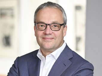 Jens Beckert Person trägt einen dunkelblauen Anzug und ein weißes Hemd, steht vor einem hellen Hintergrund mit unscharfen Bildern an der Wand.