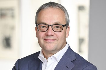 Person trägt einen dunkelblauen Anzug und ein weißes Hemd, steht vor einem hellen Hintergrund mit unscharfen Bildern an der Wand.