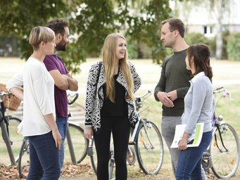 IMPRS-SPCE-Doktorandenprogramm Eine Gruppe von fünf Personen steht im Grünen, vertieft in ein Gespräch, während mehrere Fahrräder im Hintergrund zu sehen sind.