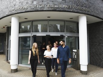 Neu am MPIfG Das Foto stellt den Eingangsbereich des MPIfG vor. Mehrere Individuen der Gattung Homo sapiens intellectualis treten heraus, andere befinden sich noch unmittelbar an der Schließtür. Sie zeigen sich einander freundlich zugewandt. Offenbar haben sie den Entschluss gefasst, gemeinsam einen Gang hinaus zu unternehmen.